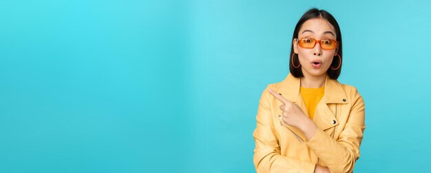 Image of asian woman looks intrigued asks question about item or store points finger left with surprised face expression stands over blue background