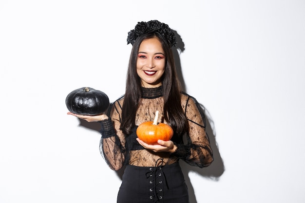 Free photo image of attractive korean girl celebrating halloween in gothic lace dress, impersonating witch and holding two pumpkins.