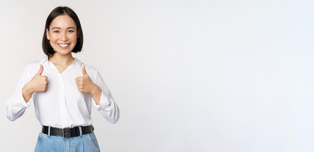 Image of beautiful adult asian woman showing thumbs up wearing formal office university clothing recommending company standing over white background