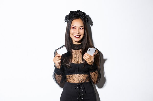 Image of beautiful asian woman in gothic lace dress and black wreath, looking aside pleased and smiling, holding mobile phone with credit card