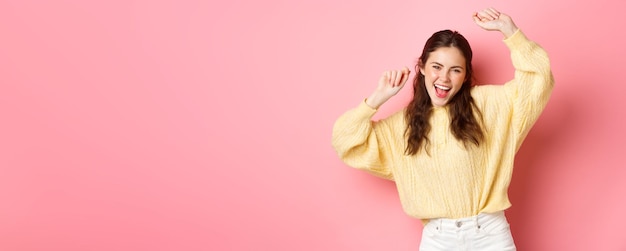 Free photo image of beautiful caucasian woman having fun dancing and enjoying party raising hands up in relaxed carefree pose standing against pink background