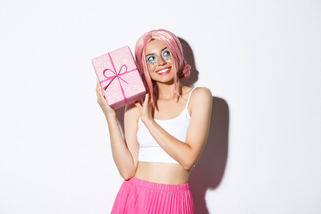 Image of beautiful girl in pink wig shaking box with birthday gift, wonder what inside wrapped box, standing.