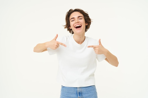 Image of brunette female model pointing at herself and smiling isolated against white studio