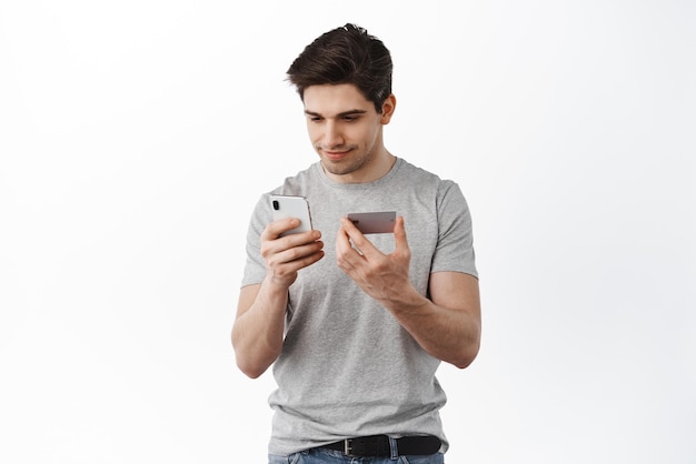 Free photo image of brunette man looking at smartphone holding plastic credit card transfer money with app paying in internet shopping standing over white background