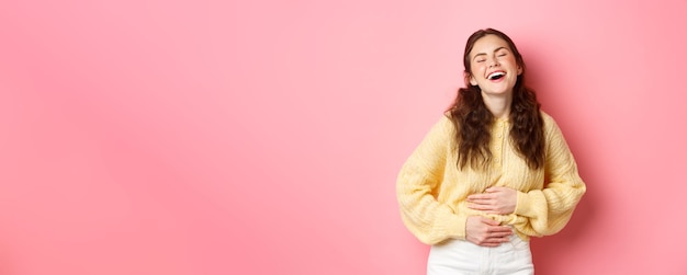 Image of carefree beautiful girl laughing out loud touching her belly while chuckling over funny comedy hilarious joke standing against pink background