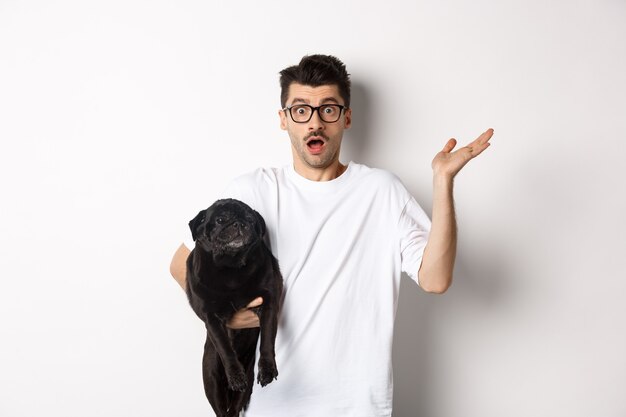 Image of confused hipster man holding dog and shrugging, dont know, raising hand puzzled, standing with his animal over white background