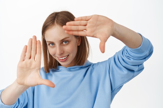 Image of creative young woman looking through hand frames gesture with pleased thoughtful smile, creating perfect shot, searching right angle, standing over white background.