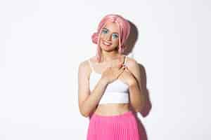 Free photo image of cute party girl in pink wig looking thankful, holding hands on heart and smiling satisfied, standing against white.