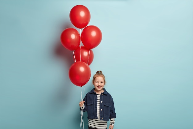 Free Photo image of cute small kid in fashionable denim jacket stands with red balloons, comes on friends birthday, has happy facial expression, stands over blue wall. childhood and celebration concept