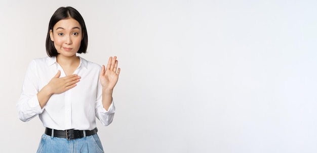 Image of cute young female office worker asian girl student raising hand up and put palm on chest name herself introduce making promise standing over white background