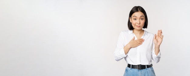 Image of cute young female office worker asian girl student raising hand up and put palm on chest name herself introduce making promise standing over white background