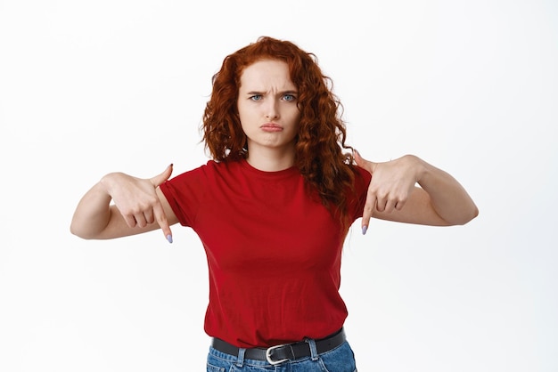 Free Photo image of disappointed and sad woman complain on unfair thing sulking and frowning pointing fingers down at banner standing against white background