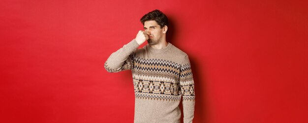 Image of disgusted young man in winter sweater shut nose and grimacing from bad smell standing over