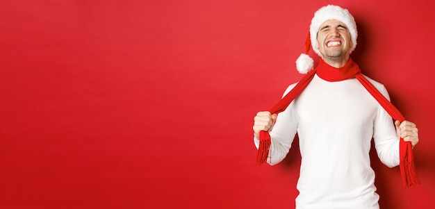 Free photo image of distressed and frustrated man in santa hat, chocking himself with scarf from sadness, standing against red background.