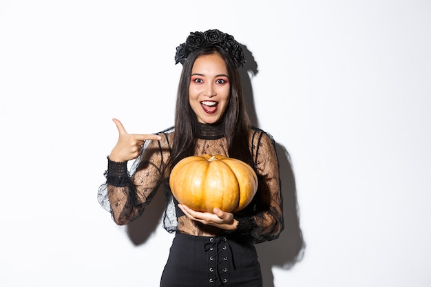 Free photo image of excited asian woman with gothic makeup, wearing black witch dress and holding pumpkin, standing amazed over white wall