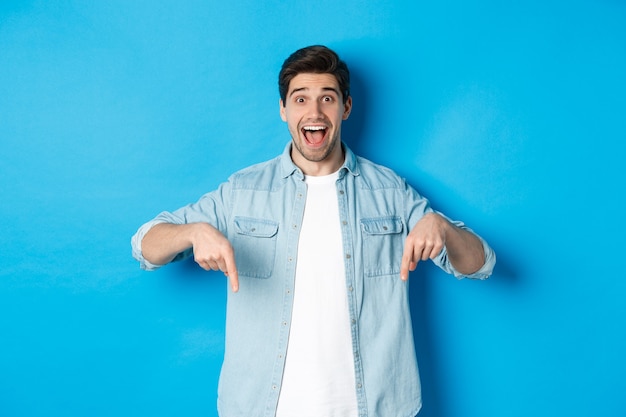 Free Photo image of excited handsome man pointing fingers down, making an announcement, standing against blue background