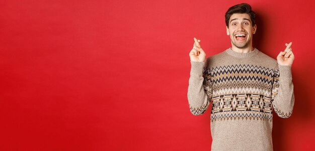 Image of excited and hopeful young man making wish wearing winter sweater crossing fingers for good ...