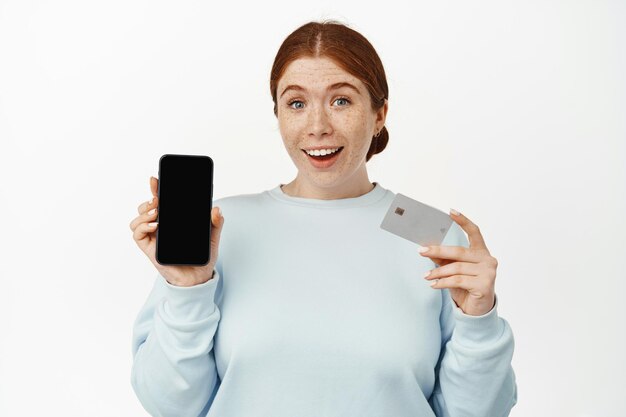 Image of excited smiling ginger girl shows her phone screen, empty mobile display and bank or discount credit card, standing in casual clothing against white background.