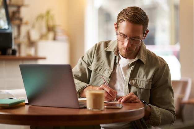 Image of focused young man in glasses sitting in cafe making notes studying attending online course