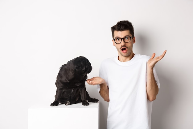 Image of funny black pug looking at his confused owner, man shrugging puzzled, standing over white background