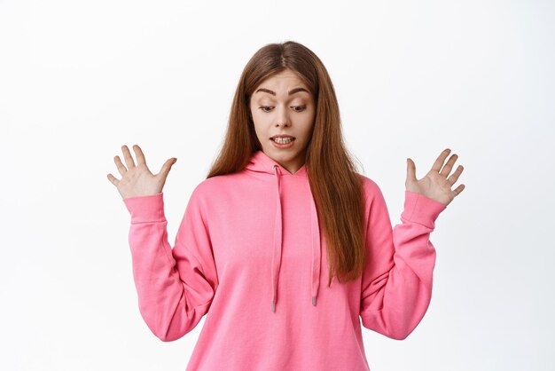 Image of girl looks surprised down spread hands sideways drop something staring at bottom standing over white background