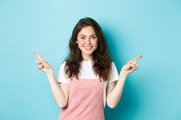 Image of glamour beautiful girl with bright make up, pointing fingers sideways and smiling, showing two variants of promo offers, give choices, blue background.