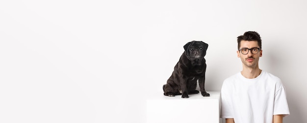 Free photo image of handsome hipster in glasses sitting next to black cute pug dog both staring at camera over