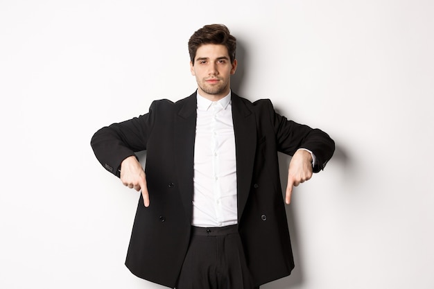 Free Photo image of handsome man dressed for formal party, wearing suit and pointing fingers down, showing advertisement or making announcement, standing over white background