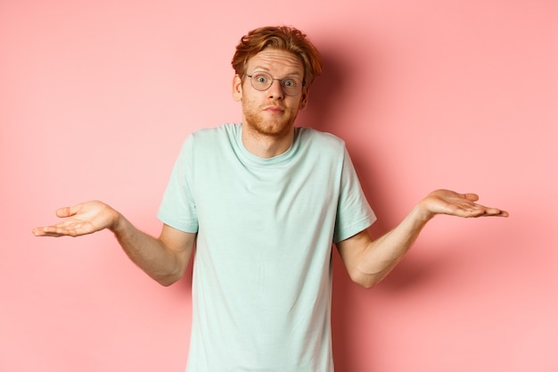 Free photo image of handsome redhead man in glasses and t-shirt know nothing, shrugging shoulders and raising eyebrows confused, standing clueless against pink background.