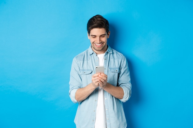 Image of handsome young man using mobile phone, texting on phone and looking pleased, standing over blue background.