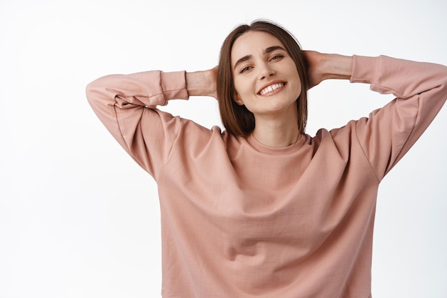 Image of happy carefree woman resting, holds hands behind head and smiling satisfied, feeling joy and freedom, enjoy weekend holiday, standing against white background