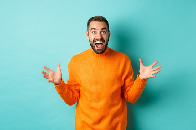 Free photo image of happy and excited man announce big news, spread hands and shouting of joy, rejoicing, standing over light turquoise wall.