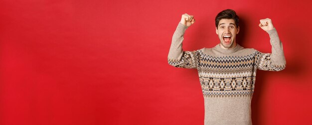 Image of happy handsome guy rejoicing on christmas eve raising hands up and shouting for joy yes achieve new year goal standing over red background in sweater