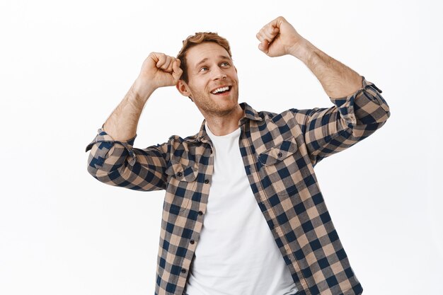 Image of happy redhead man dancing and looking at upper right corner promotional text, having fun, partying and doing success victory dance, white wall