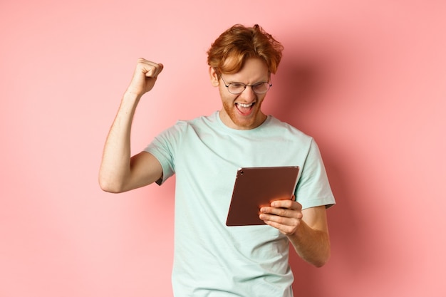 Image of happy redhead man triumphing winning online with digital tablet and rejoicing standing over...