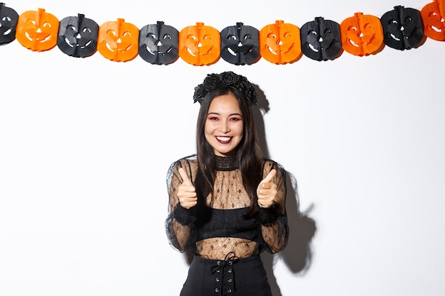 Free photo image of happy and satisfied asian female in witch costume showing super, thumbs-up gesture and smiling pleased, standing against pumpkins banner.