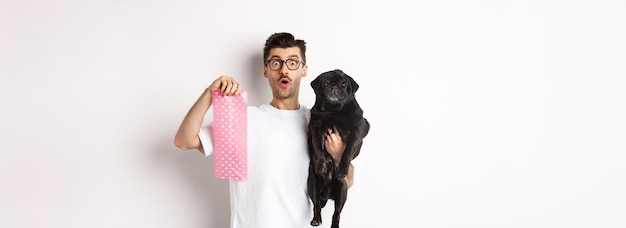 Free Photo image of hipster guy pet owner holding cute black pug and dog poop bag standing over white backgroun