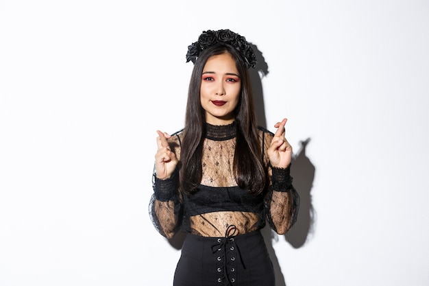 Image of hopeful asian girl in gothic lace dress and wreath making wish, cross fingers for good luck, looking wishful at camera, standing over white background.