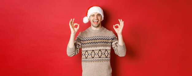 Image of joyful attractive guy in sweater and santa hat wishing merry christmas showing okay signs a