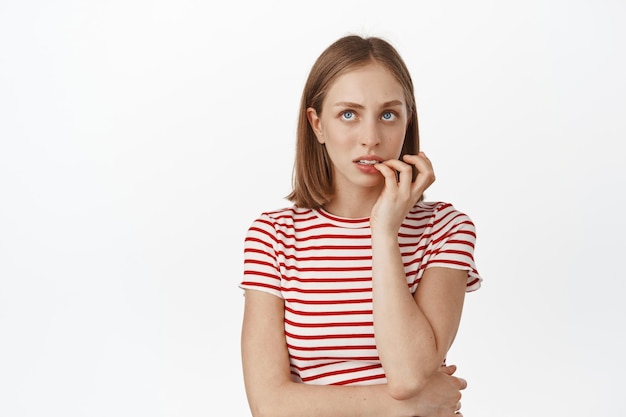 Image of nervous girl student, blond woman biting finger and looking up with concerned thoughtful face, thinking, having anxiety, standing against white background