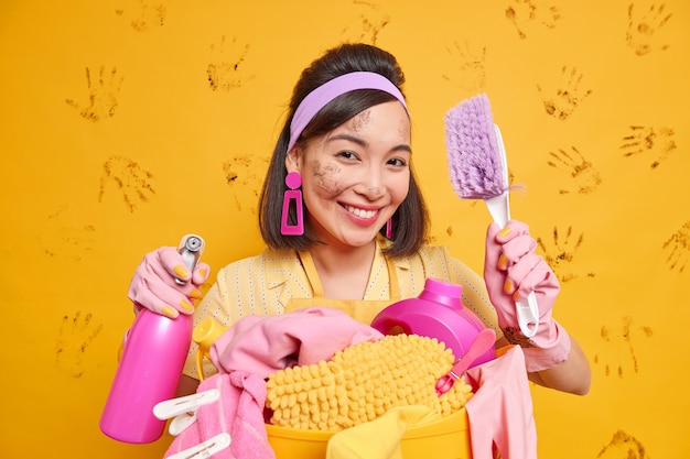 Free photo image of positive housewife smiles pleasantly holds cleaning tools enjoys working process poses with brush and detergent poses near basket of laundry poses against yellow wall