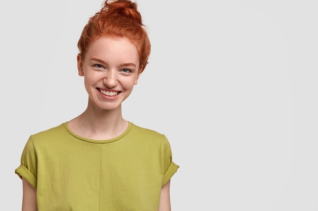 Free photo image of pretty red haired girl has charming smile on face, wears casual green t shirt, feels happy, isolated over white wall with free space for your advertising content or promotion. emotions