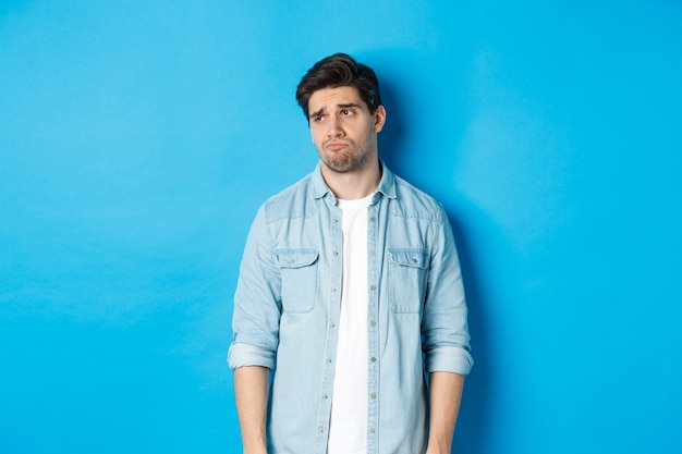 Free photo image of reluctant and sad guy in casual outfit looking left, frowning and feeling upset, standing against blue background