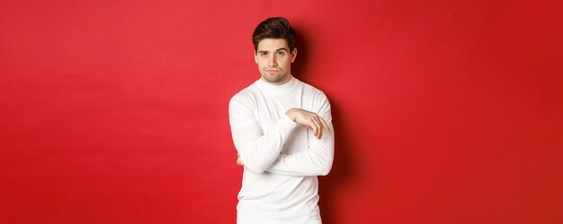 Image of sassy caucasian man in white sweater looking thoughtful at camera making choice standing ov...