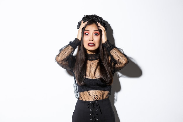 Free photo image of scared and worried asian woman in halloween costume looking horrified or anxious, holding hands on head and stare at camera, panicking over white background.