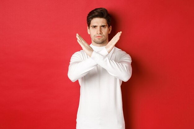 Image of serious and angry man in white sweater express storng disapproval showing cross sign to sto...