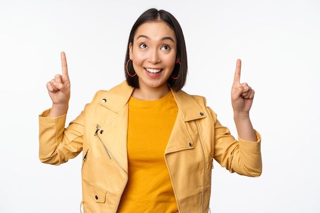 Image of smiling asian brunette woman pointing fingers up showing advertisement with happy face posing against white background