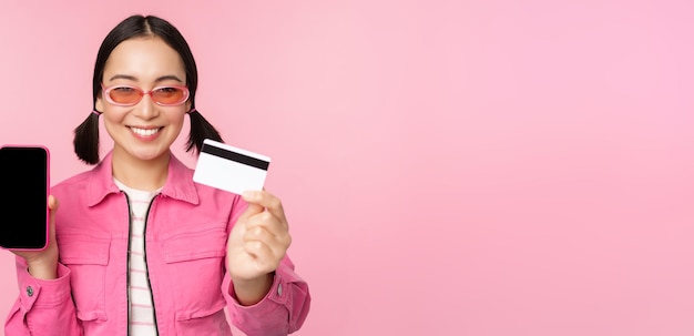 Free photo image of smiling korean woman showing credit card and mobile phone screen smartphone application interface paying online shopping contactless standing over pink background