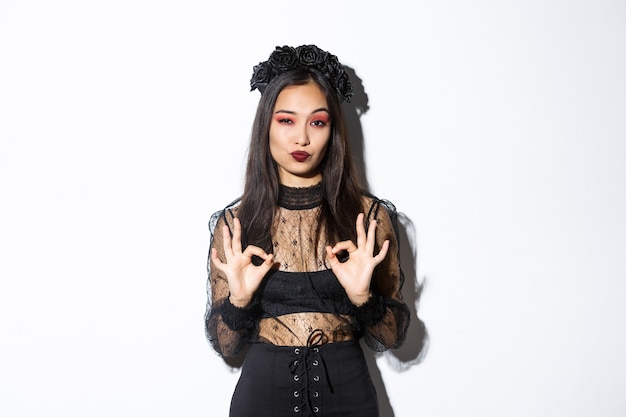 Free photo image of stylish asian woman in gothic dress, looking satisfied and showing okay gestures in approval, praise something good, standing impressed over white background.