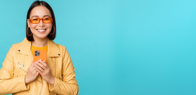 Image of stylish beautiful chinese woman in sunglasses holding smartphone taking photo on mobile phone and smiling recording smth standing over blue background